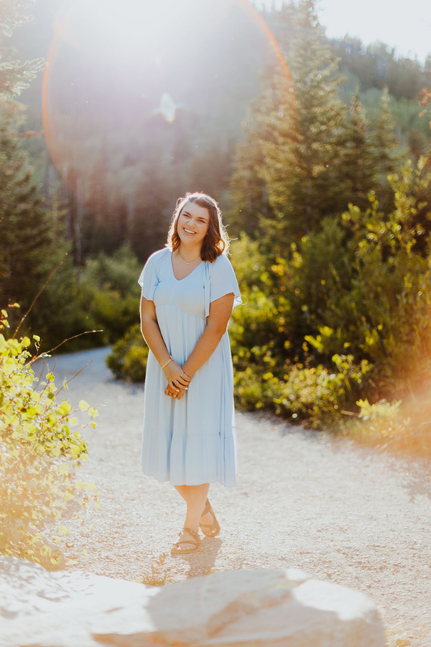 Light-filled portrait of someone in a blue dress standing on a mountain trail surrounded by evergreen trees at sunset.