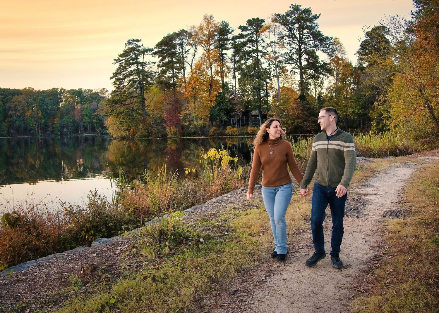 A couple walking along a scenic waterfront trail during golden hour with tall pine trees.