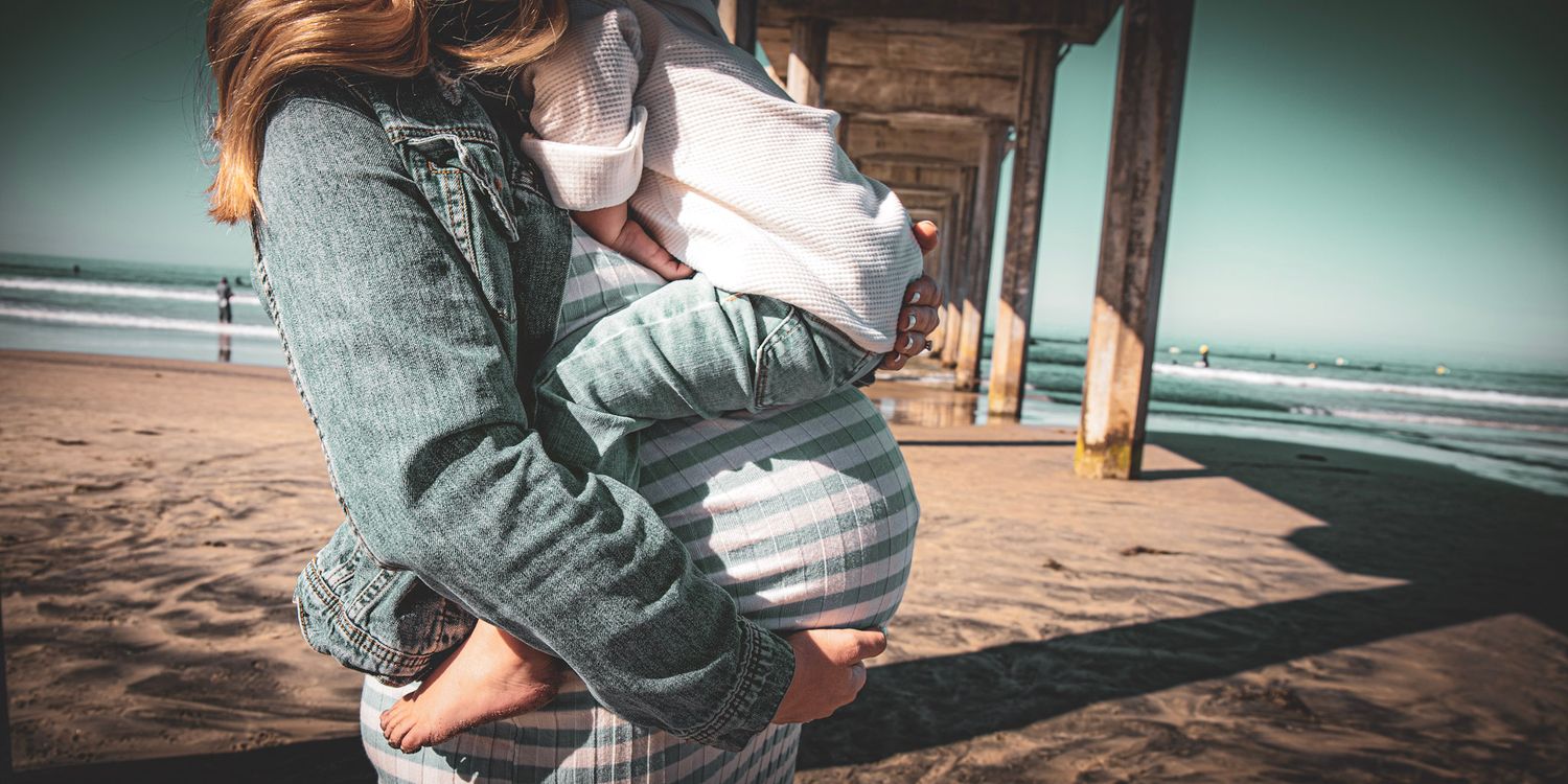 Low angle shot of figure holding baby against beach pier backdrop.