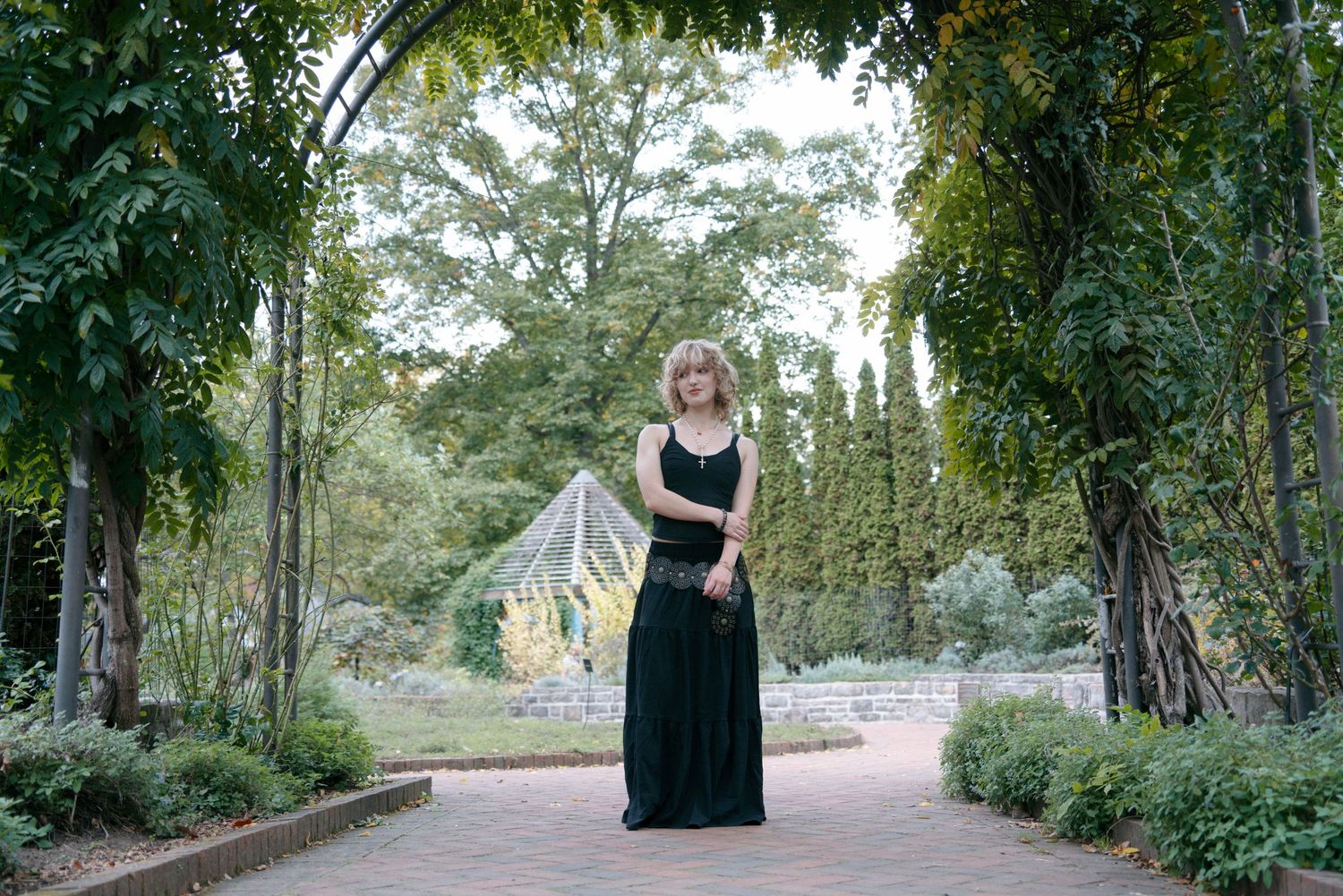 Woman in elegant green maxi dress poses under a garden archway surrounded by lush greenery.