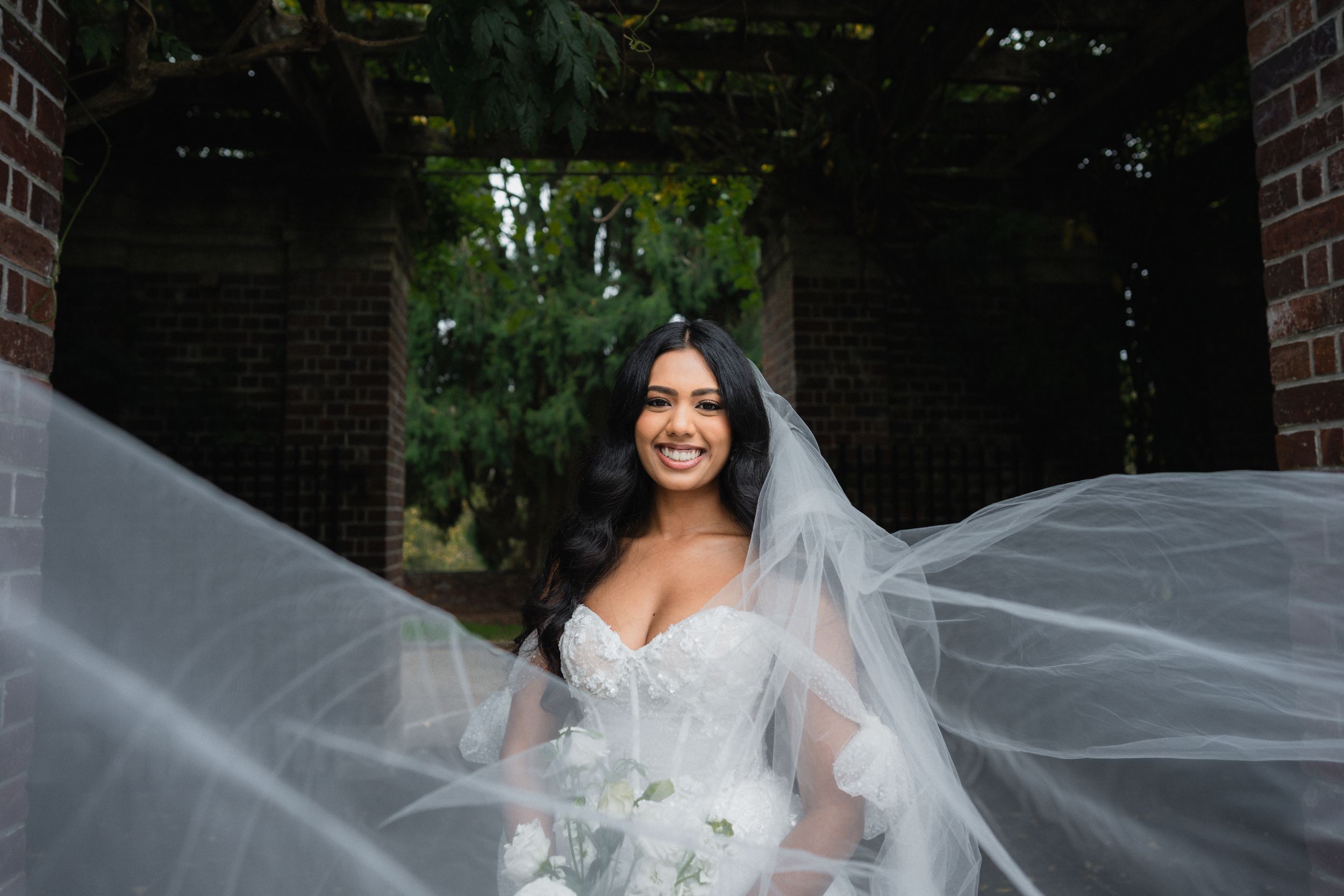 A bride in a white wedding dress with flowing veil poses outdoors against a brick backdrop.