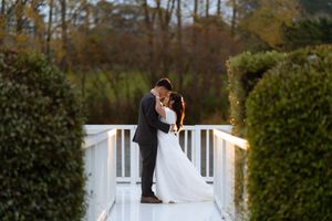 Bride and groom share a romantic kiss on a white bridge surrounded by lush hedges and autumn trees at golden hour.