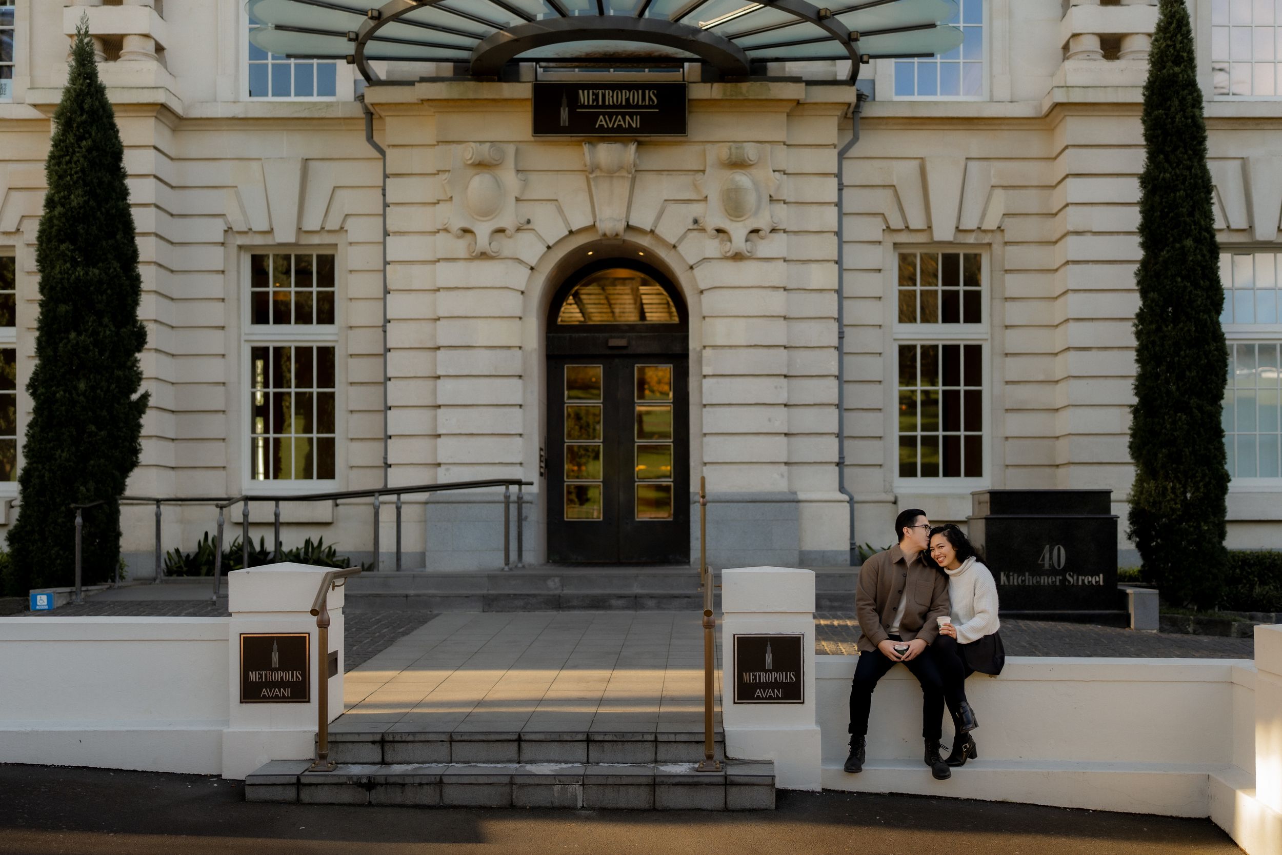 Couple sitting on steps outside a grand hotel entrance with classical architecture and tall cypress trees.