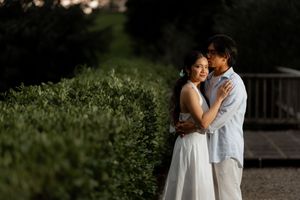 A couple embraces tenderly in a lush garden, the woman in a white dress and man in a blue shirt, bathed in soft golden light.