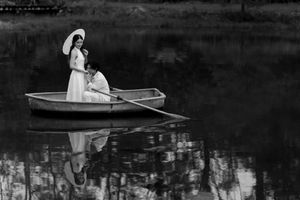 Woman in white dress sitting in rowboat on calm water with reflection in black and white photograph.