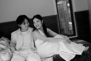 Two women sitting together on a bed in a black and white photograph, smiling at the camera in an intimate moment.