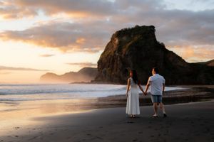 Couple holding hands on a sandy beach at sunset with dramatic rocky cliffs and ocean waves in background.