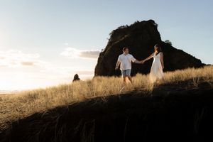 Couple holding hands walking on coastal cliffside grass at golden hour with dramatic rock formation backdrop.