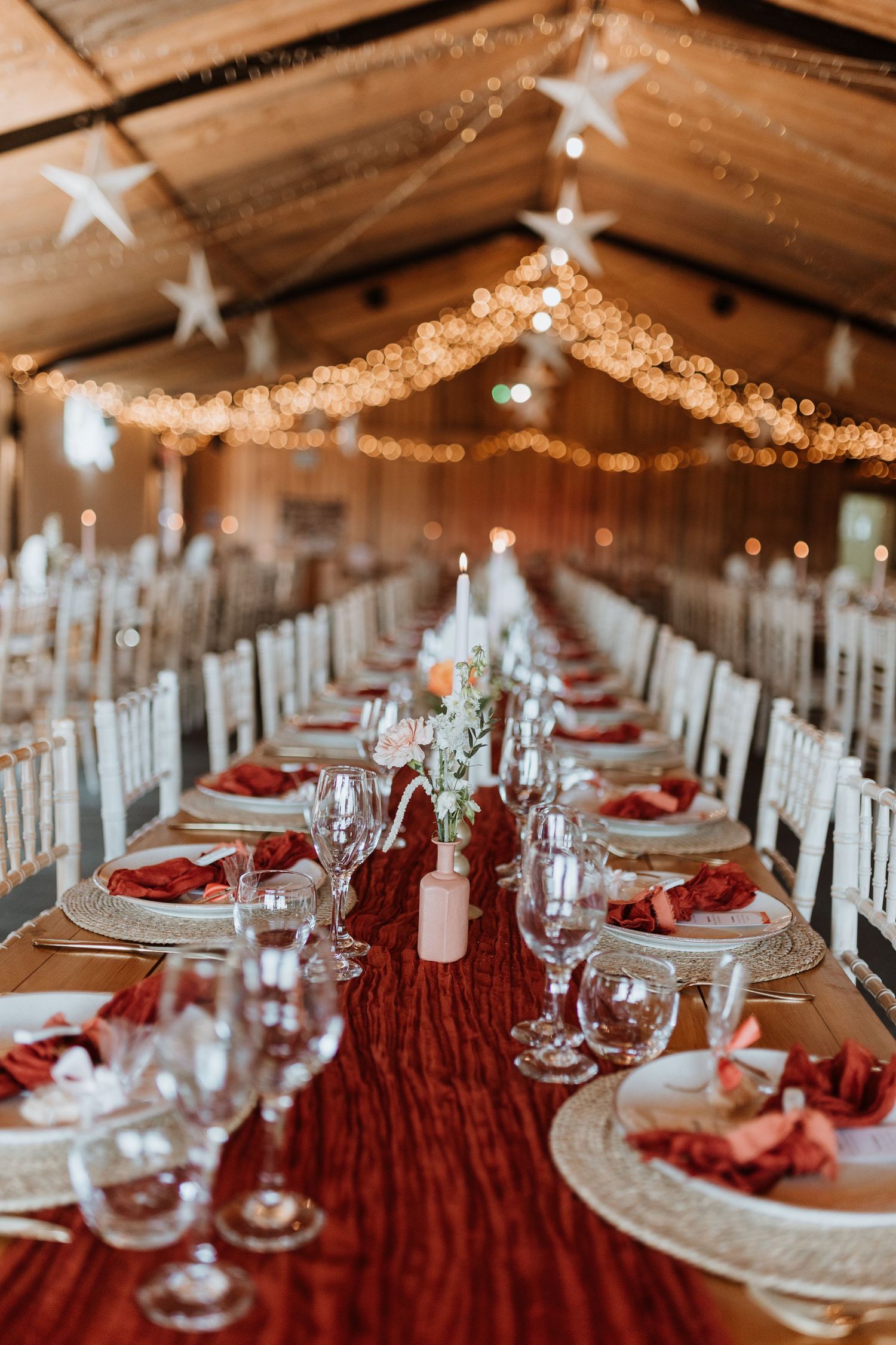 Elegant wedding reception tables set with red tablecloths, white plates and twinkling lights strung from rustic wooden ceiling.