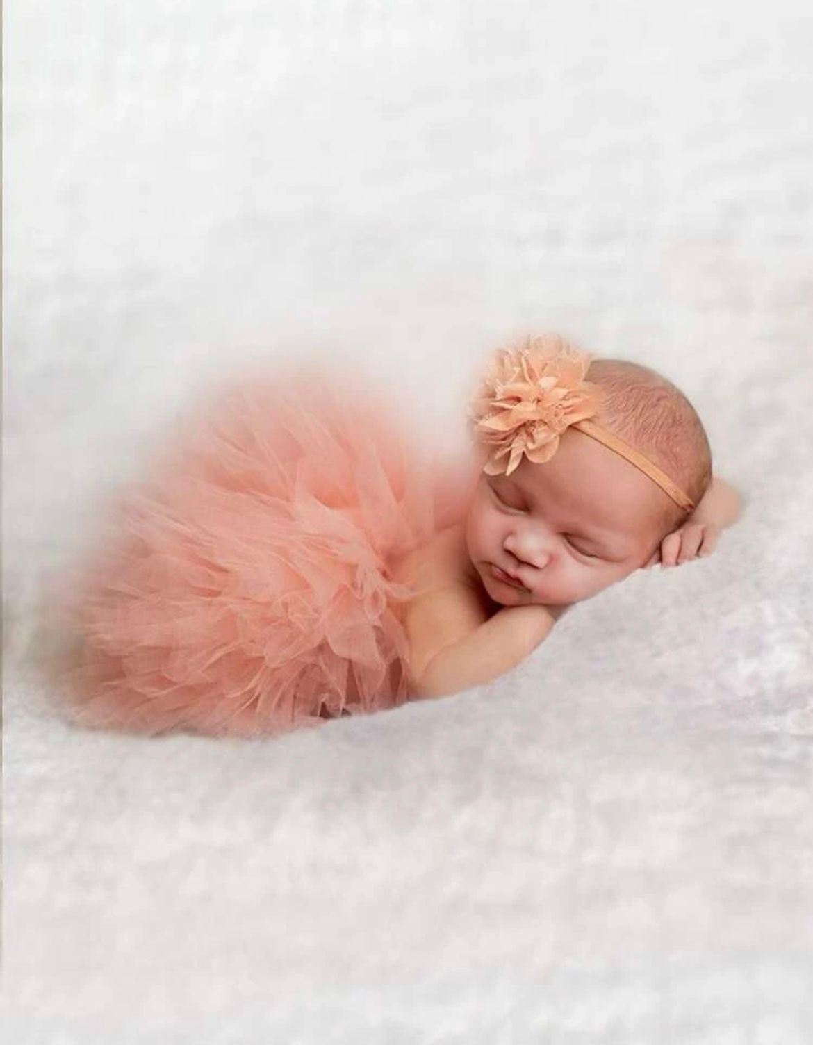 Newborn baby wearing peach tutu and headband sleeping on white blanket.