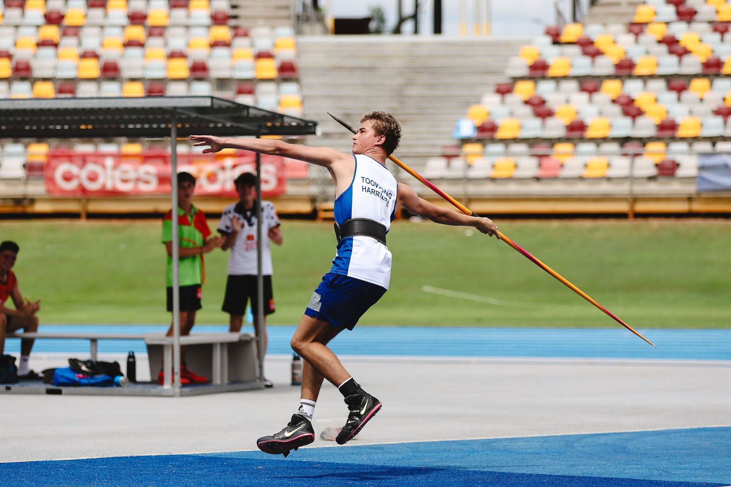 An athlete performs a javelin throw at a track and field stadium with colorful stadium seats in the background.