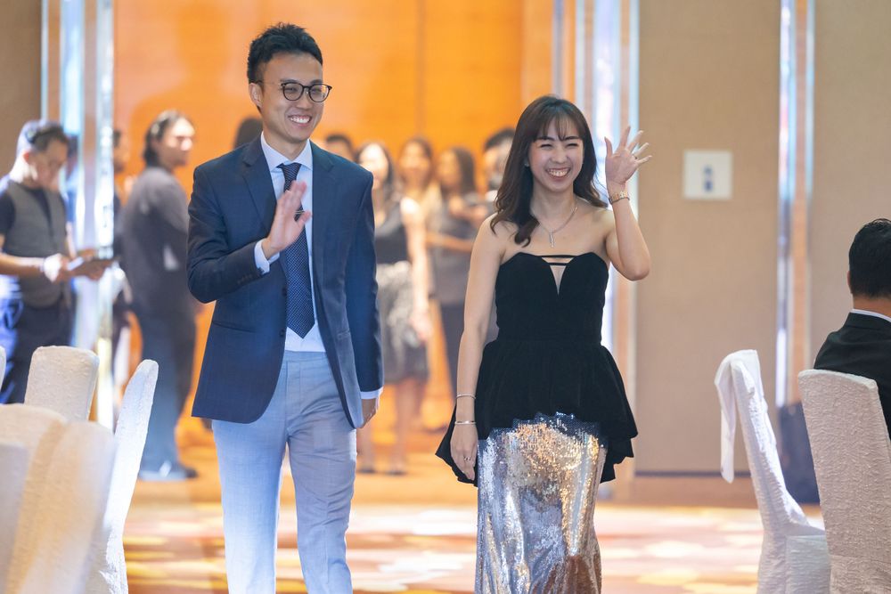Two people walk and wave in a formal event venue with gold walls and white chairs in the background.