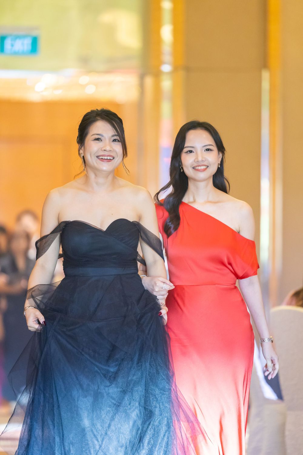 Two women in evening gowns, one wearing a black off-shoulder dress and one in a red one-shoulder dress walking together.