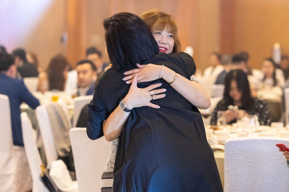 Two people share an emotional embrace at a formal event in a banquet hall filled with seated guests.