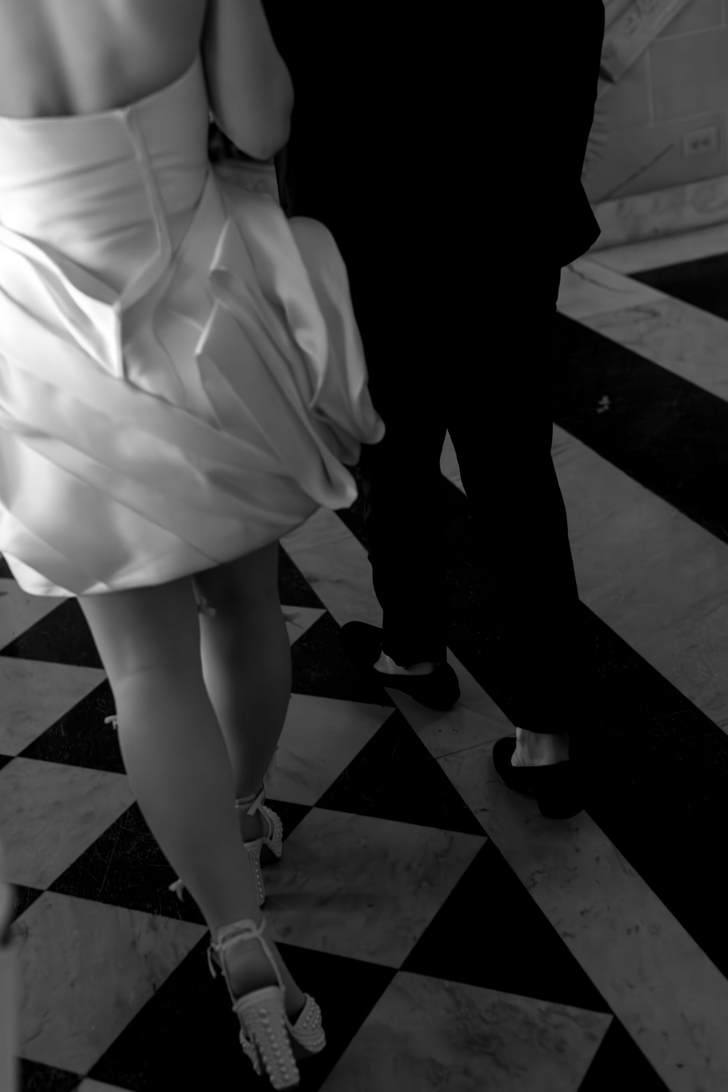 A couple dancing on a black and white checkered floor, with the woman wearing a white dress and the man in dark attire.
