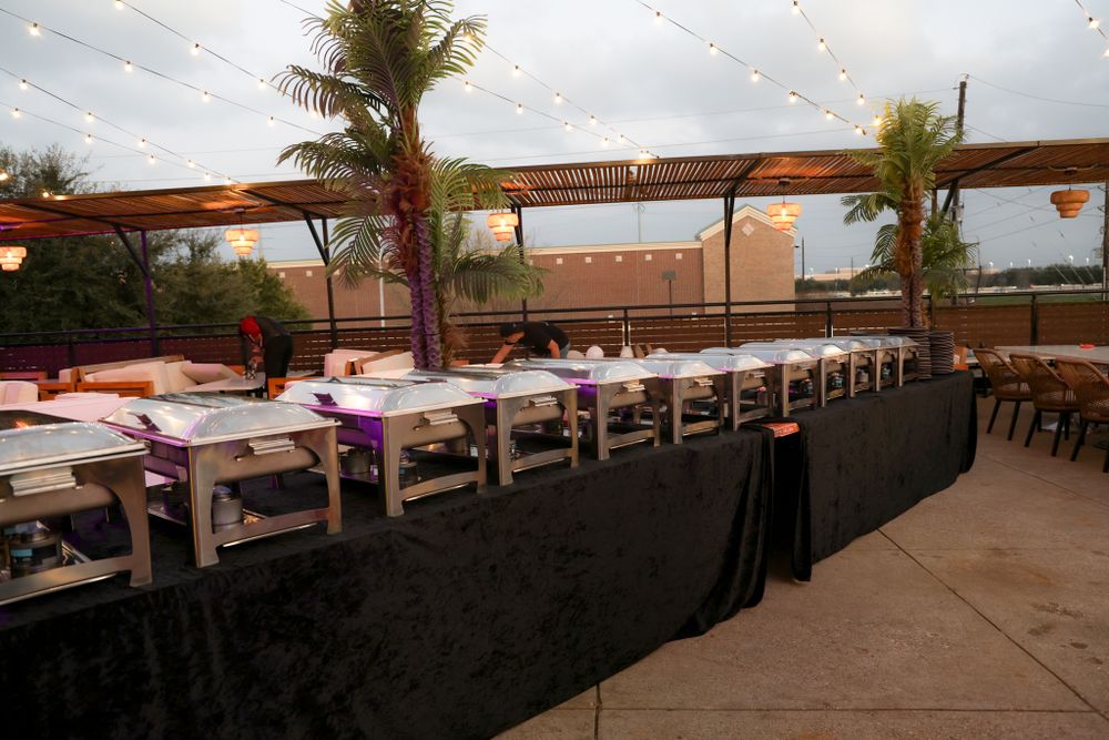 Long tables set up at an outdoor venue with palm trees and string lighting overhead.