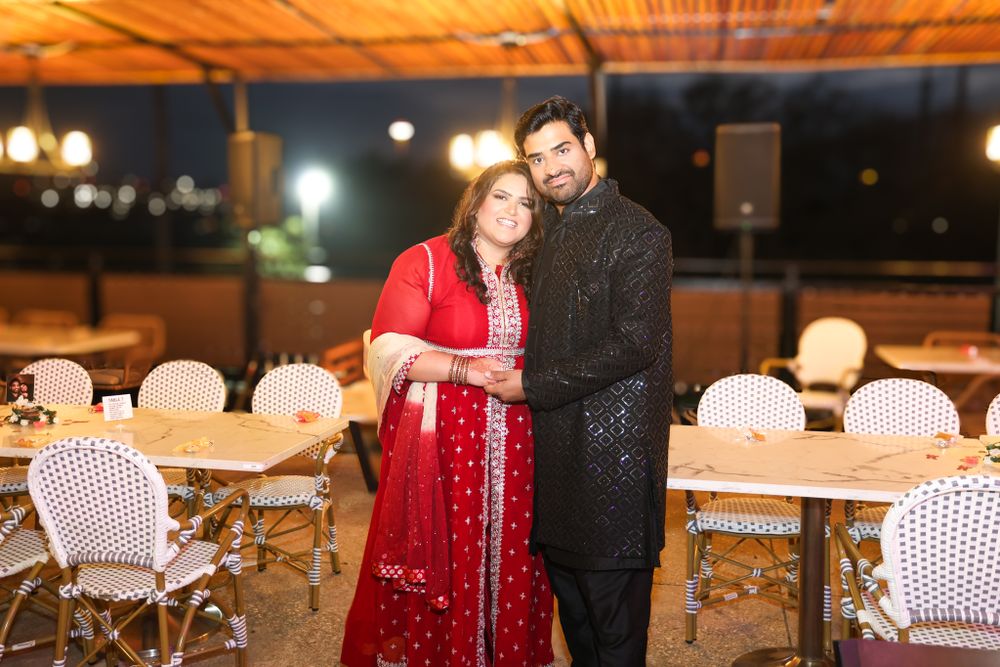 A couple in formal Indian attire stands together at an outdoor evening venue.