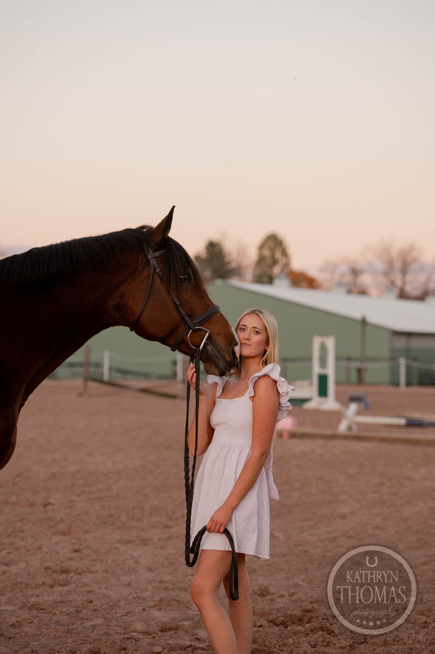 A person in a white dress stands close to a dark horse's face in an equestrian setting.
