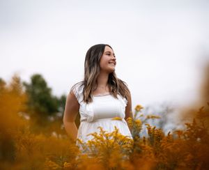 Woman in white dress stands surrounded by golden wildflowers against cloudy sky during autumn season.