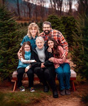 A family poses together on a wooden bench surrounded by evergreen trees at a Christmas tree farm in autumn.