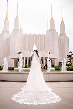 A bride in a white lace veil stands before the Washington DC Mormon Temple with fountains in the foreground.