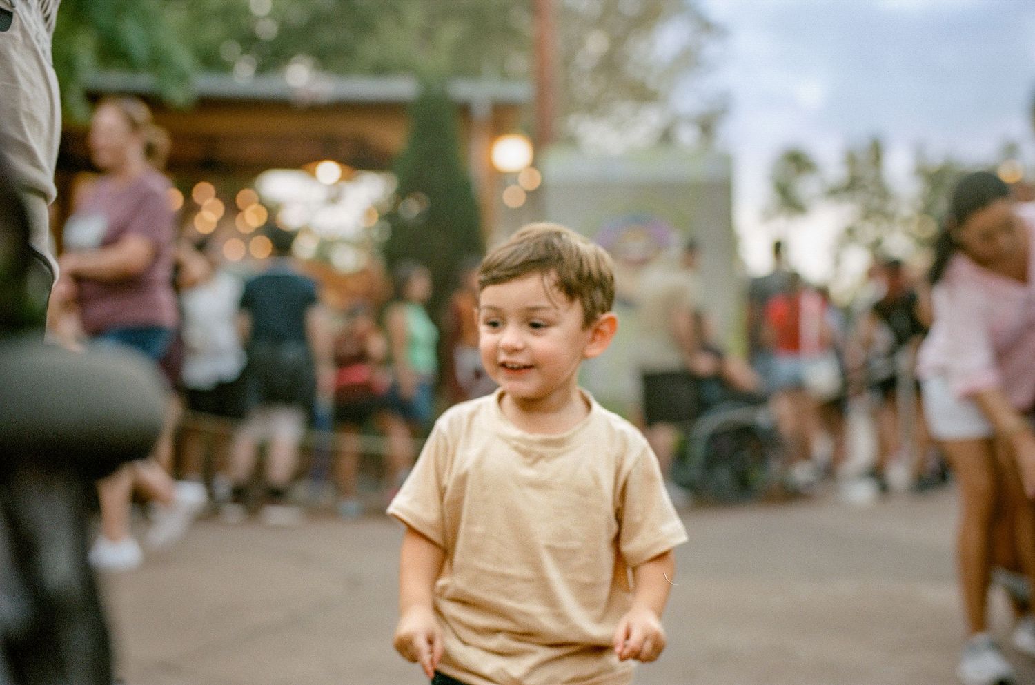A child in a beige t-shirt runs joyfully through a busy outdoor gathering at dusk.