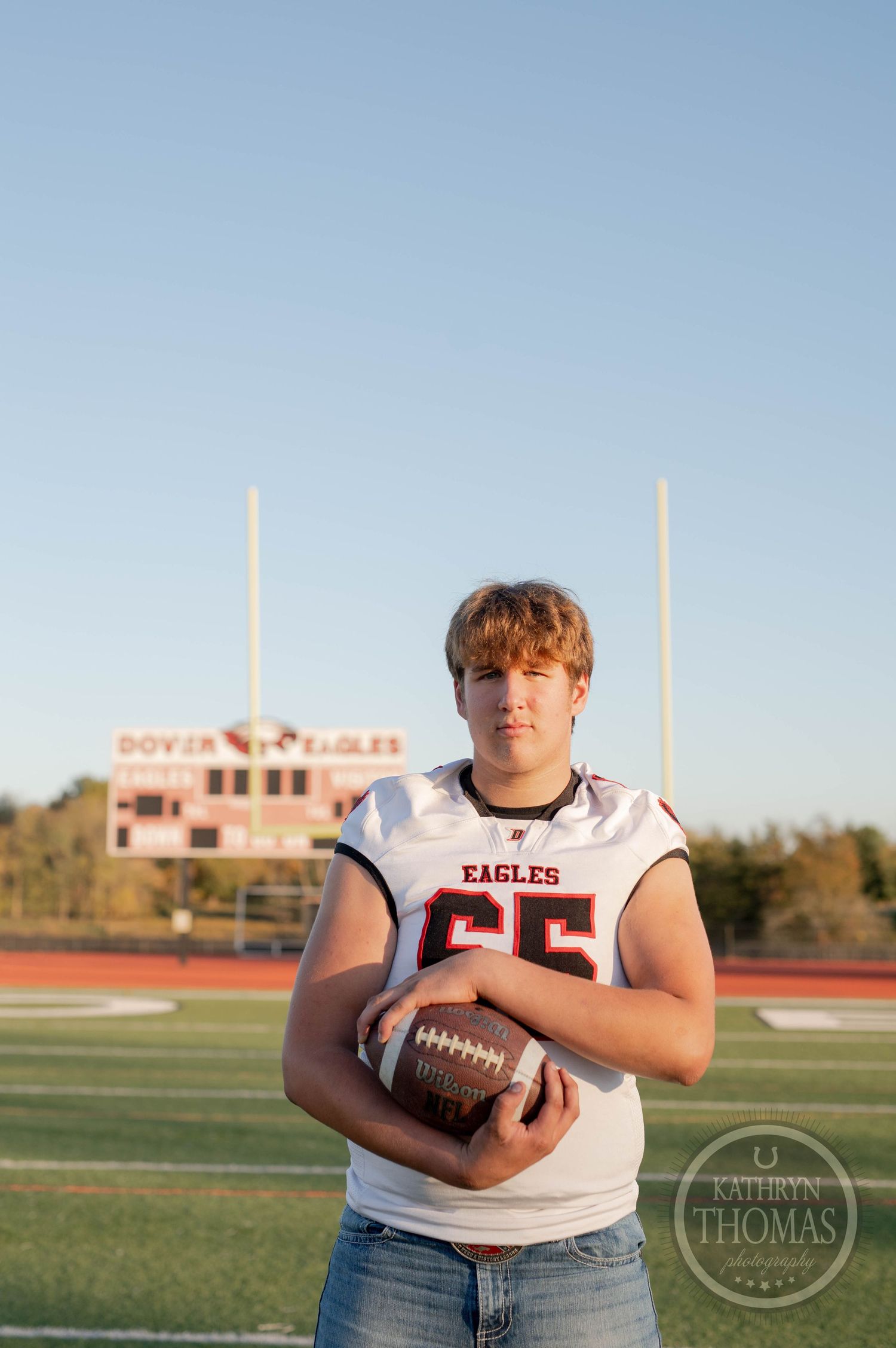 Eagles player number 65 stands on football field holding ball with goal posts and scoreboard in background.