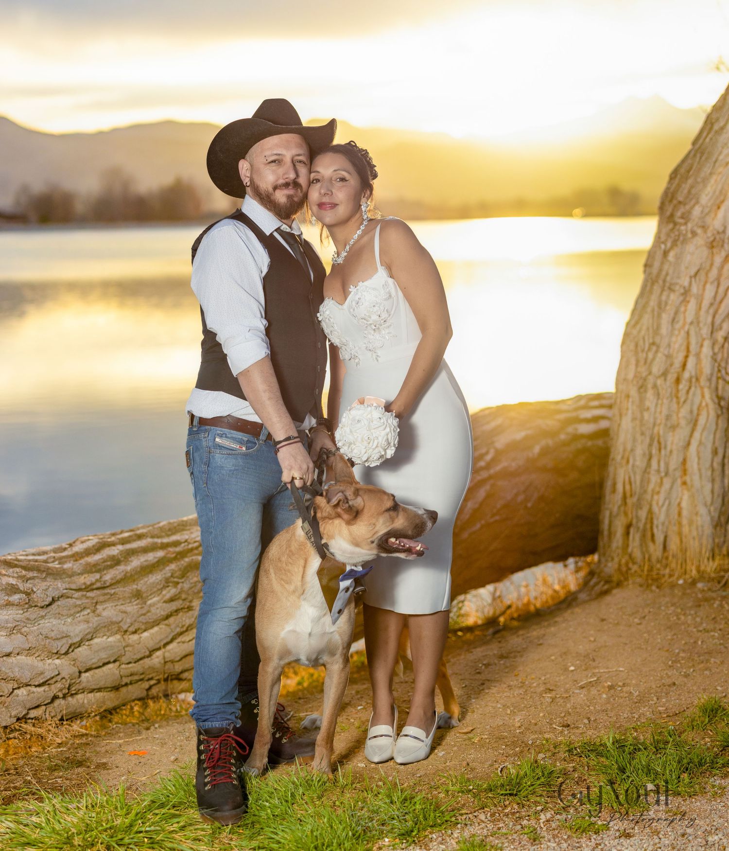 A couple and their dog pose for a romantic sunset photo by a lake with mountains in the background.