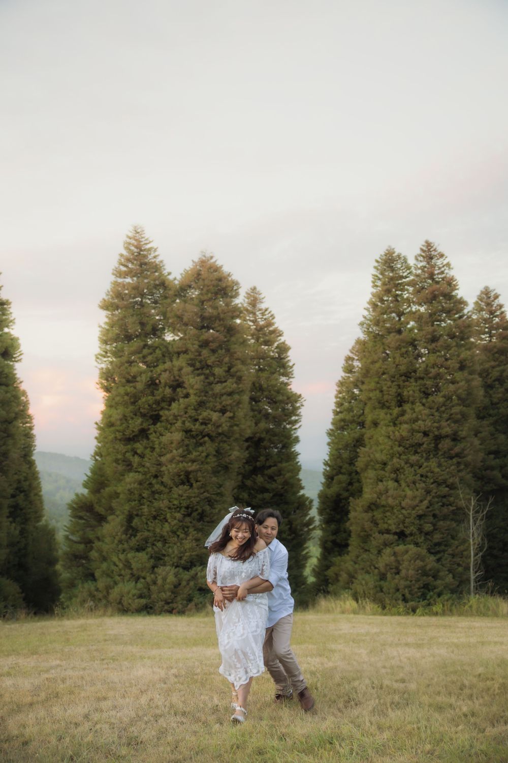 A couple dances playfully in a mountain meadow with towering evergreen trees in the background.