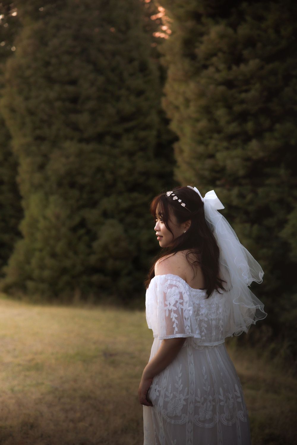 A bride in a white lace dress poses in front of evergreen trees during a sunset mountain photoshoot.