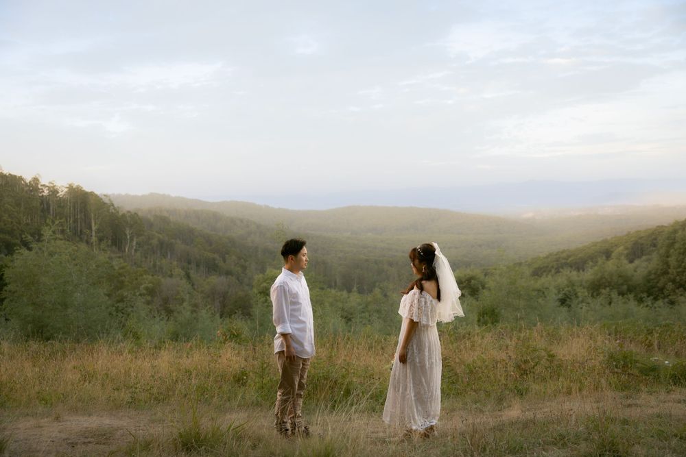 A couple stands together on a hilltop overlooking a misty valley landscape at dusk.