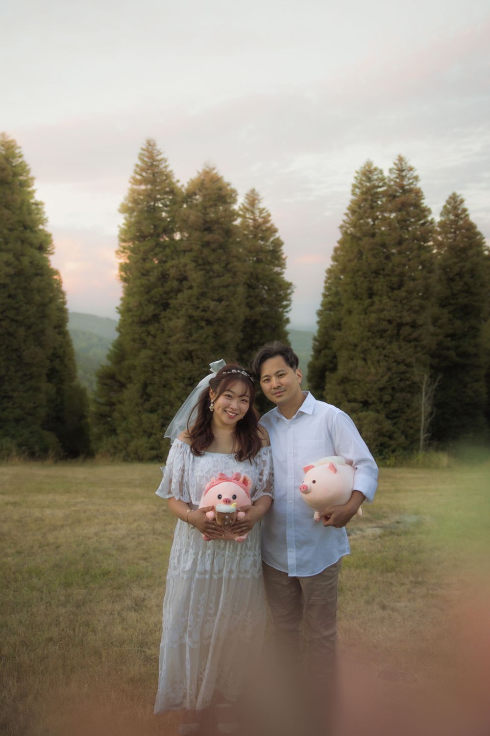A couple holding pink plush toys stands in front of tall evergreen trees at sunset.