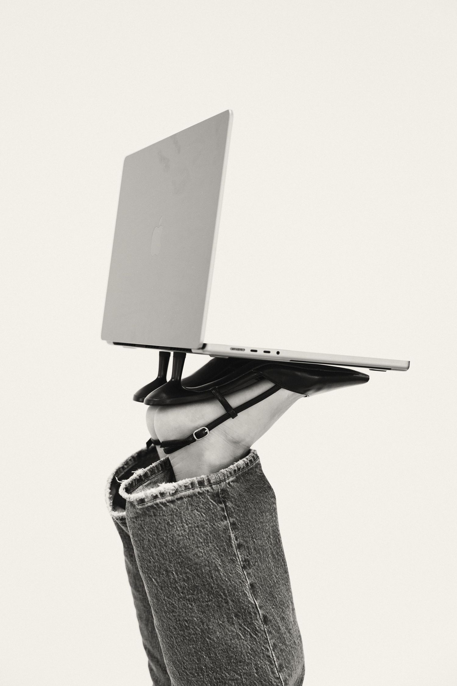 Black and white photo of person in jeans holding Apple MacBook laptop overhead against white background.