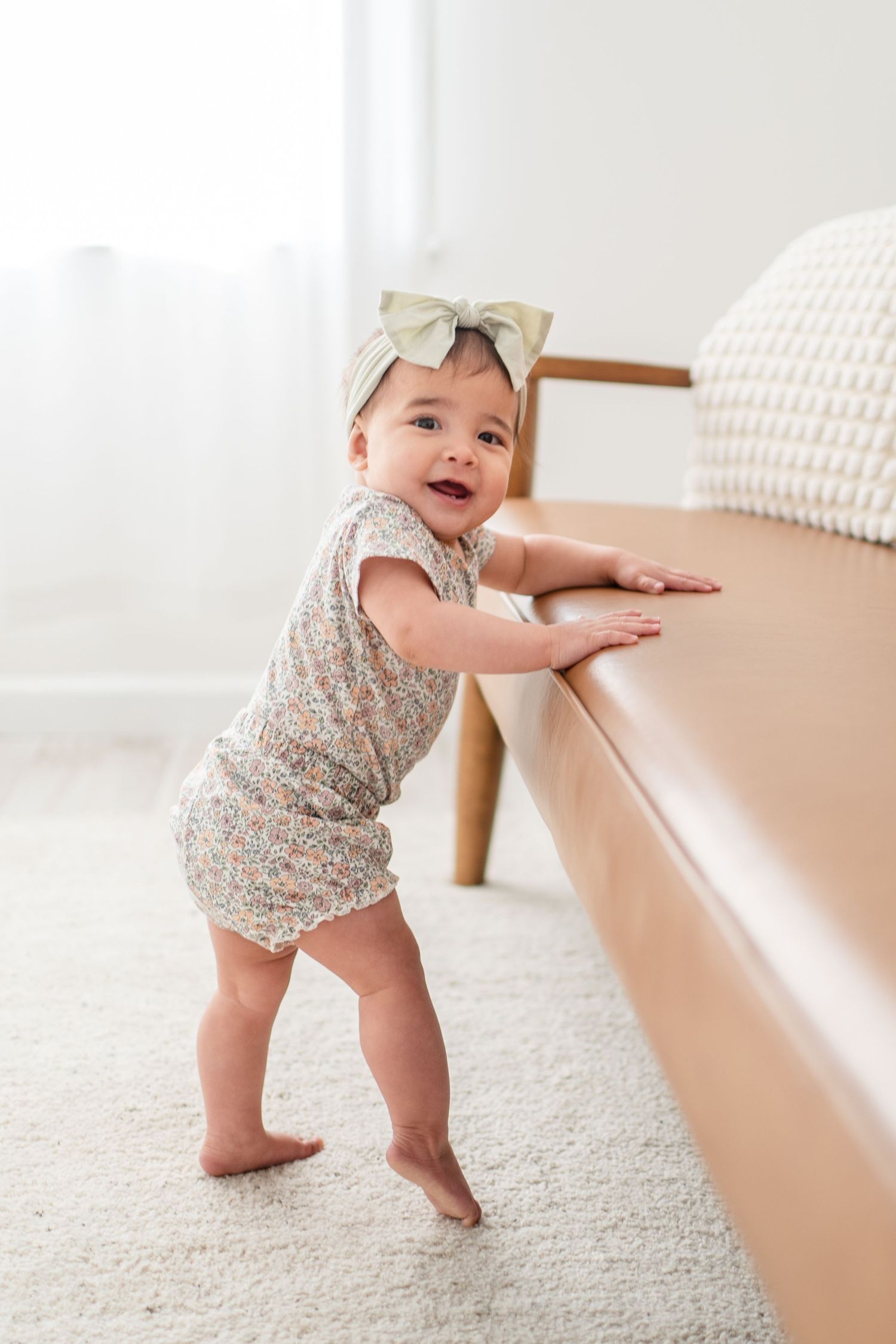 Small child in floral romper and bow headband stands at brown couch while learning to walk on carpeted floor.