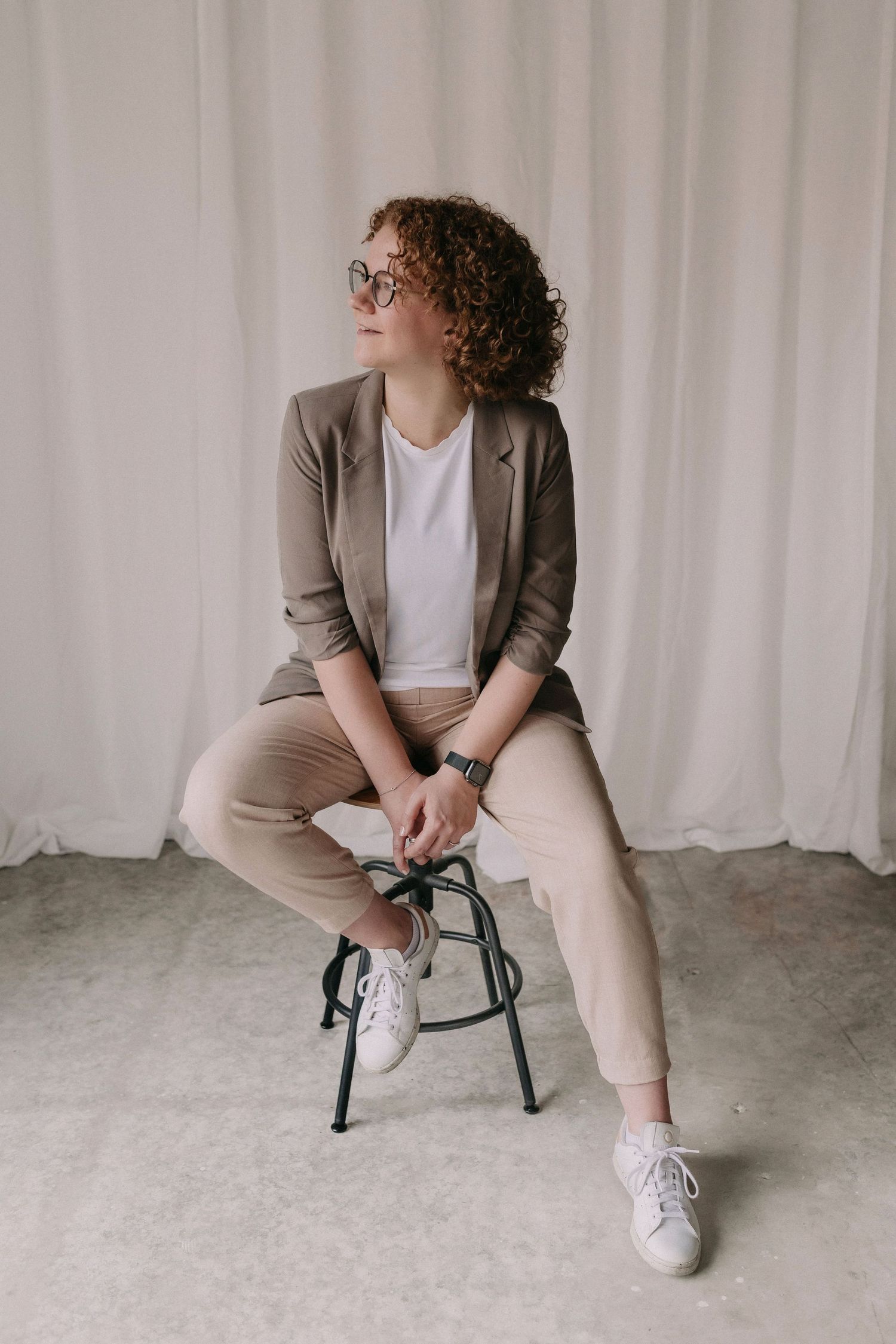Professional in business casual attire with curly hair and glasses sits on metal stool against white backdrop.