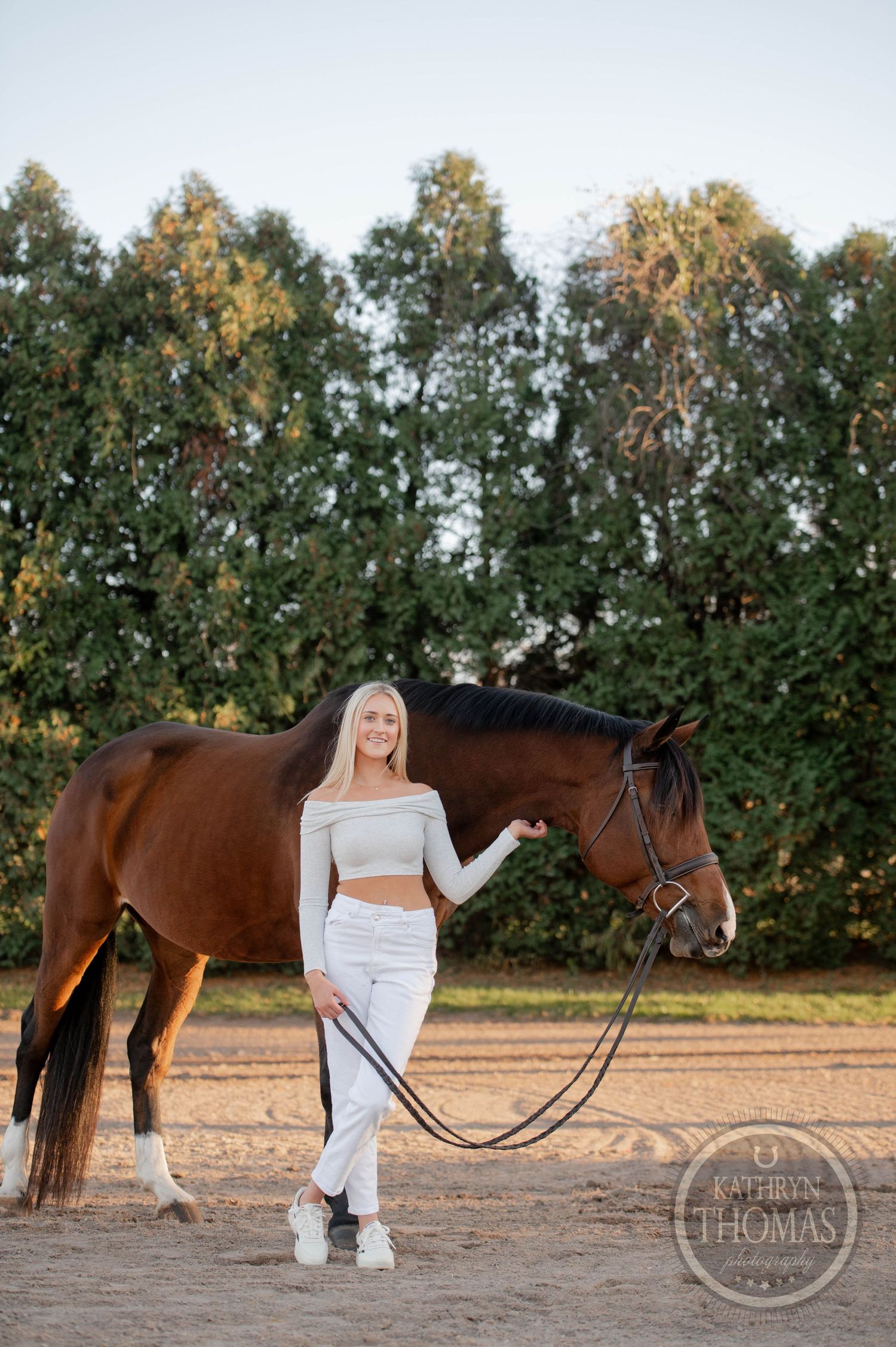 A white-clad figure stands beside a bay horse in an outdoor arena with trees in the background at sunset.