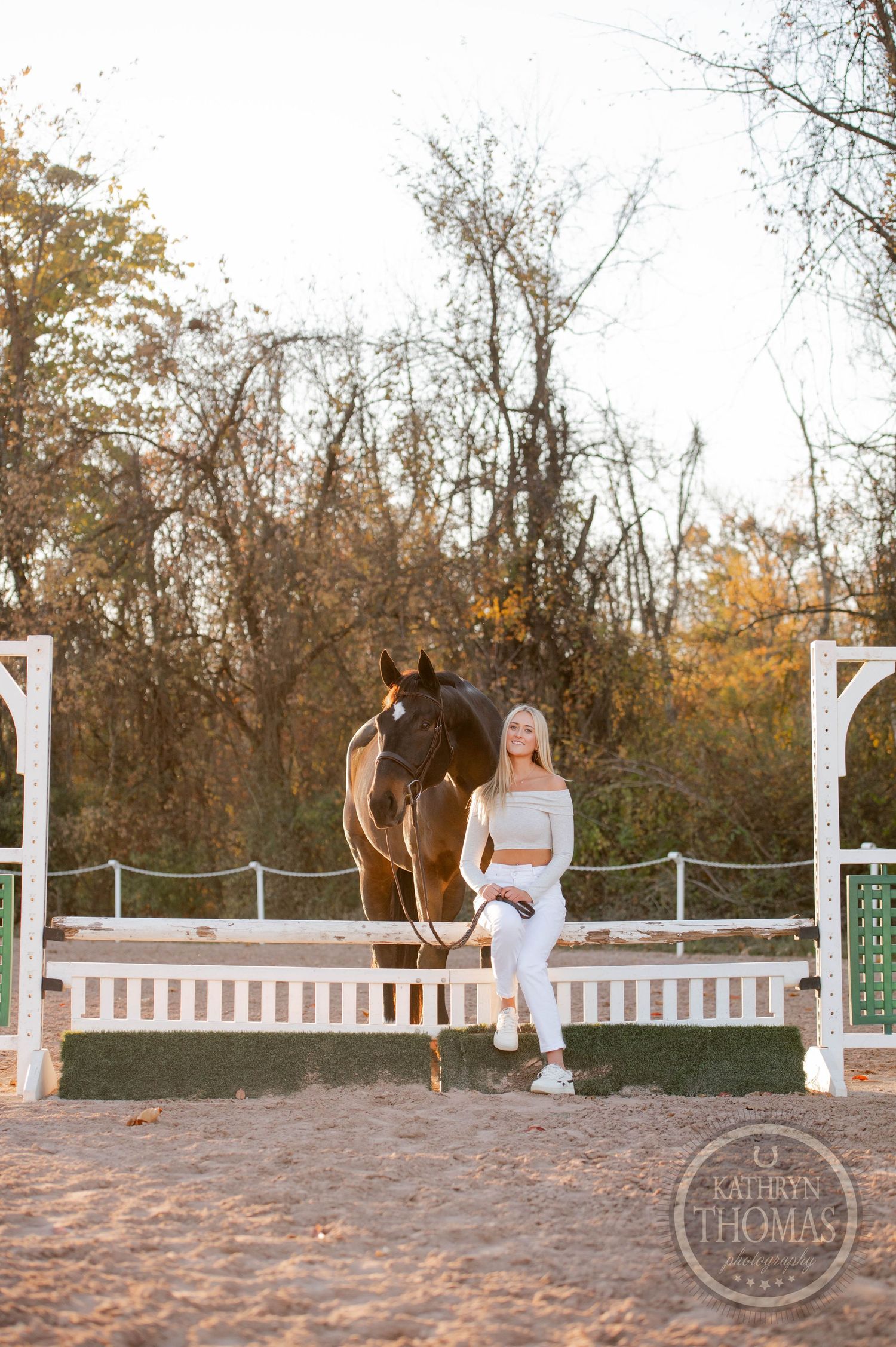 A jumping horse and someone in white attire pose by decorative white posts in an autumn setting.