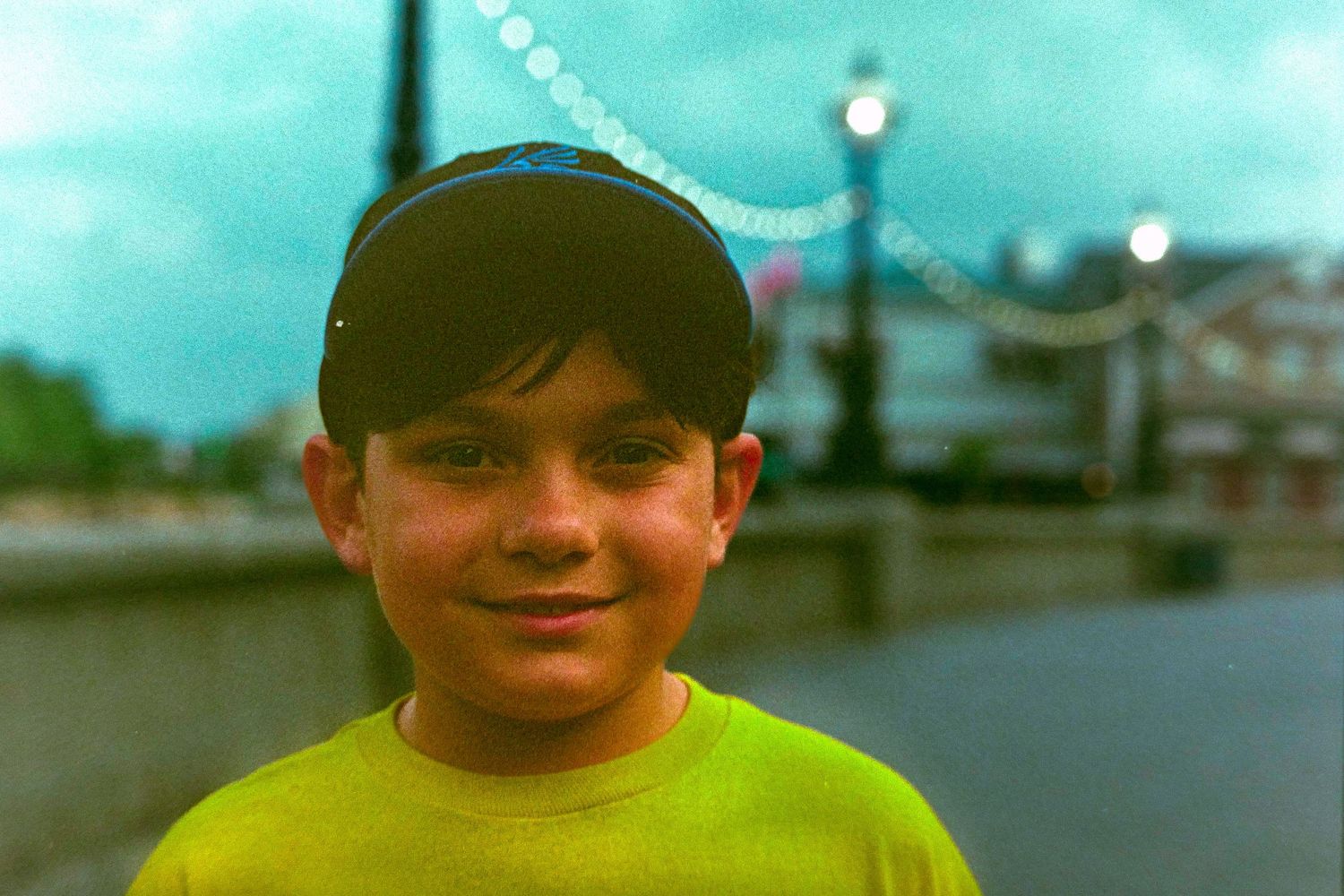 A young person in a bright yellow t-shirt and baseball cap smiles at the camera against a turquoise-tinted background.