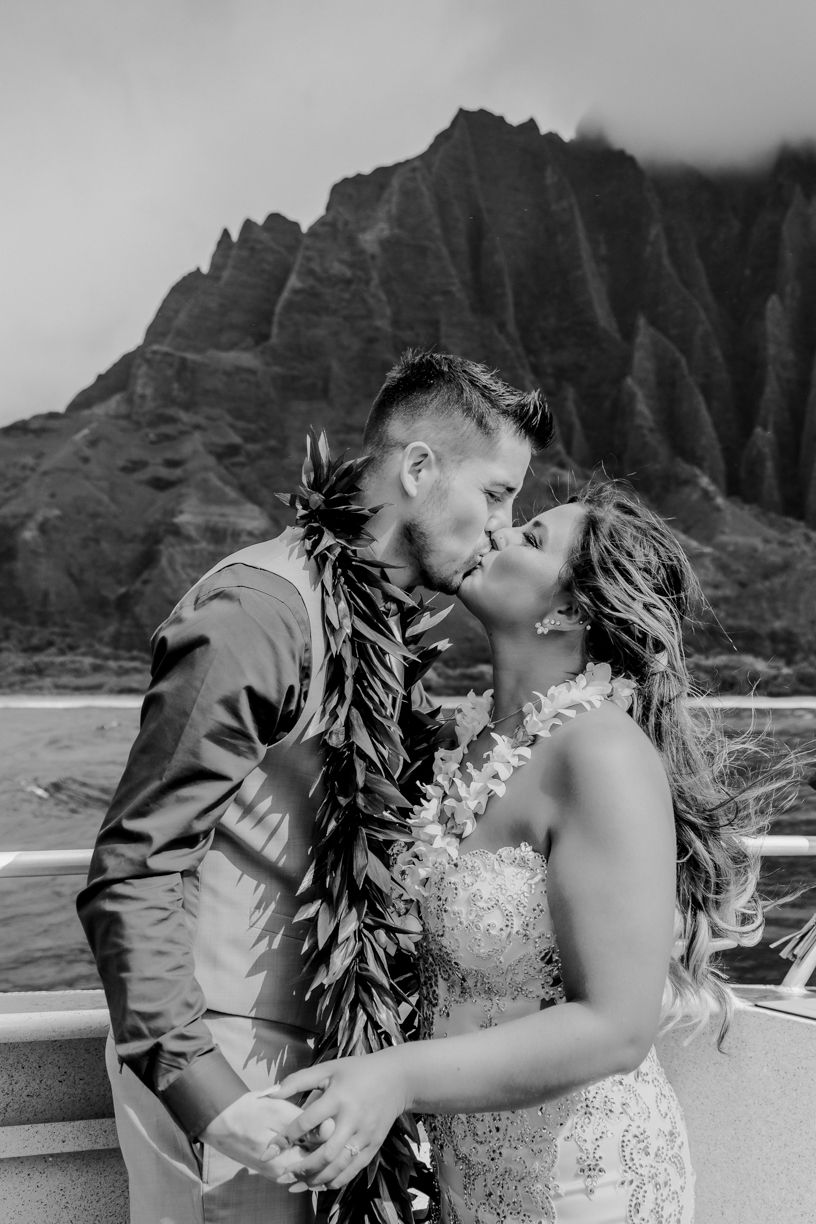 A romantic wedding couple sharing a kiss aboard a boat with dramatic Na Pali coast mountains of Kauai in the background.