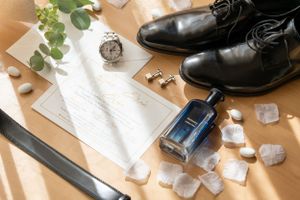 Wedding accessories including black dress shoes, perfume bottle, and rings on paper.