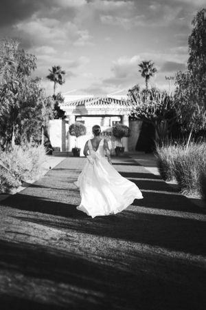 A bride in a flowing white dress twirls on a path leading to a Spanish-style villa. Next day wedding photography.