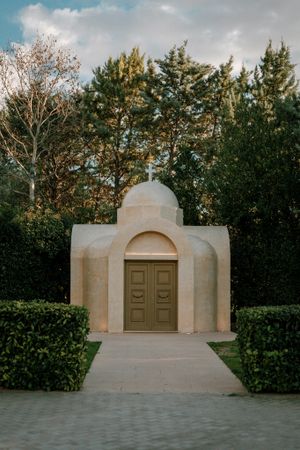 Small white chapel with arched entrance and cross on top surrounded by evergreen trees.