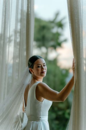 Elegant bride in simple white dress poses near sheer curtains with palm trees visible outside. Next day wedding photography.