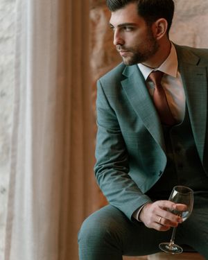 Sophisticated man in green suit holding wine glass while sitting by window. Next day wedding photography.