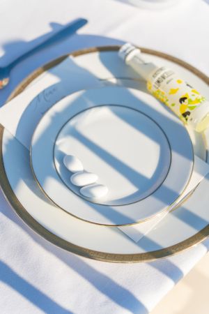 Place setting with gold-rimmed plates and striped shadows on white tablecloth.