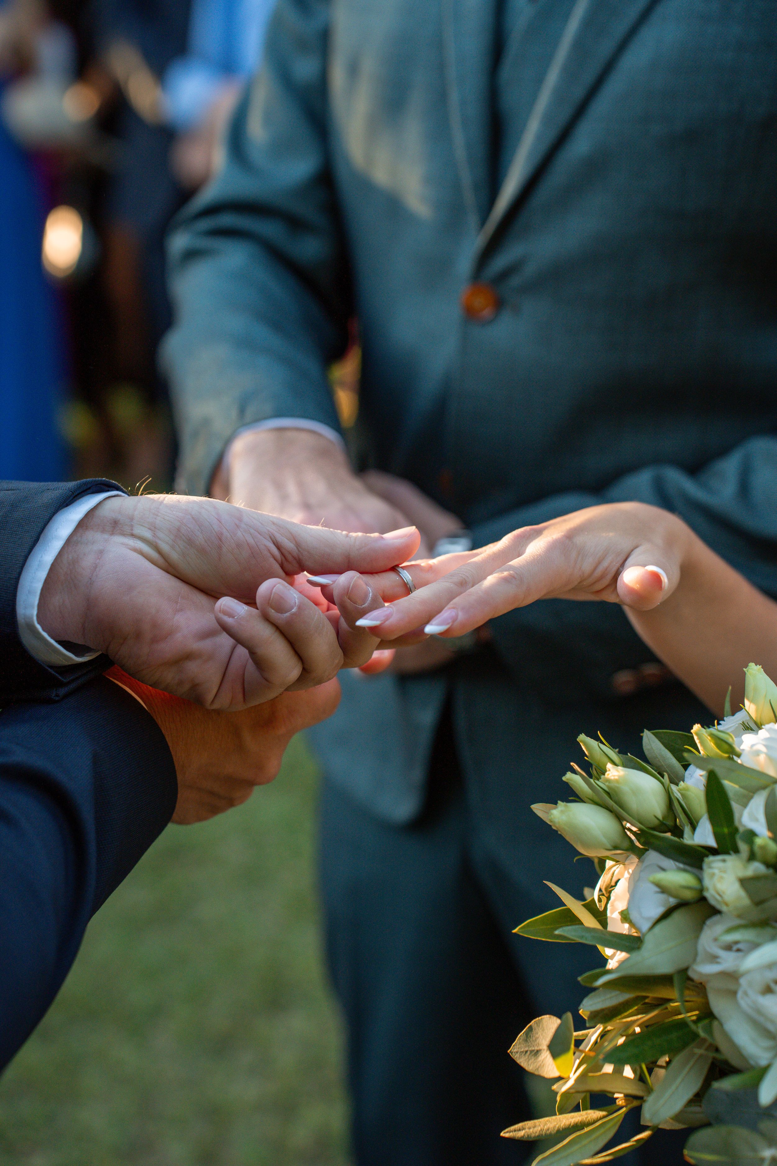 A close-up of hands exchanging wedding rings during an outdoor ceremony with a bouquet visible.