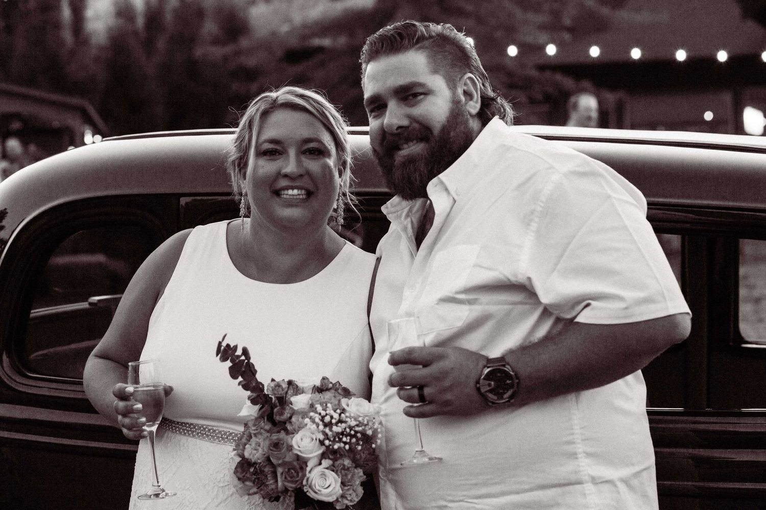 Black and white wedding photo in front of vintage car with floral bouquet and string lights overhead.