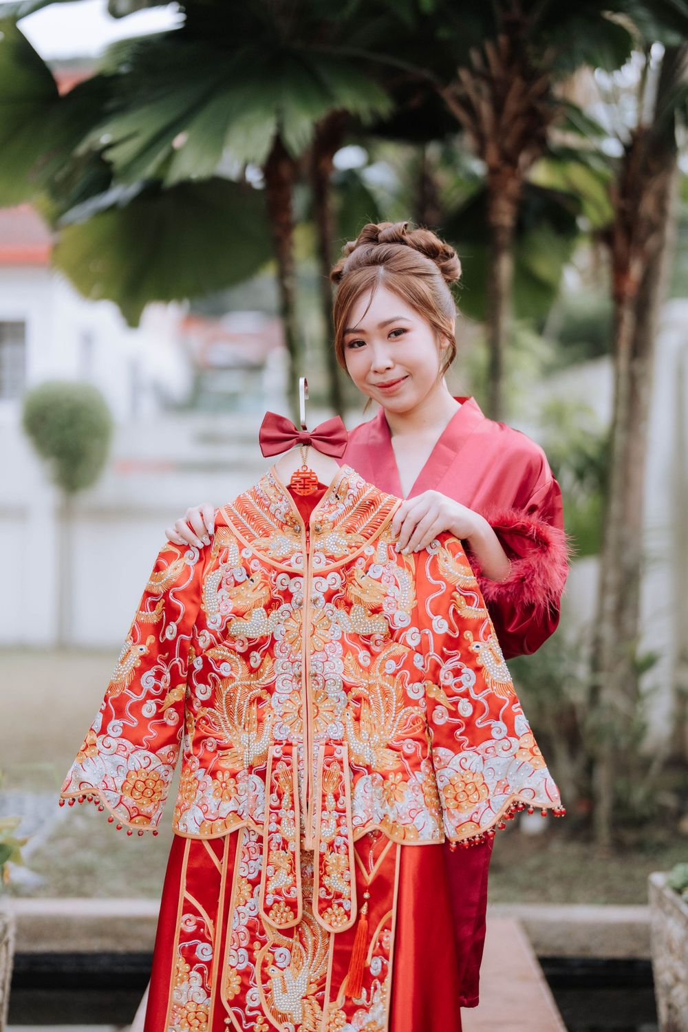 Person in red robe holding up an ornate traditional Chinese wedding dress outdoors.