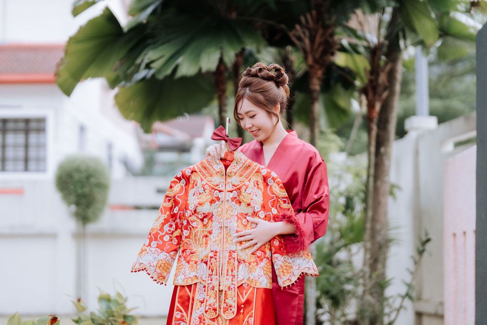 Person in red robe displaying an ornate Chinese wedding dress in a garden setting.