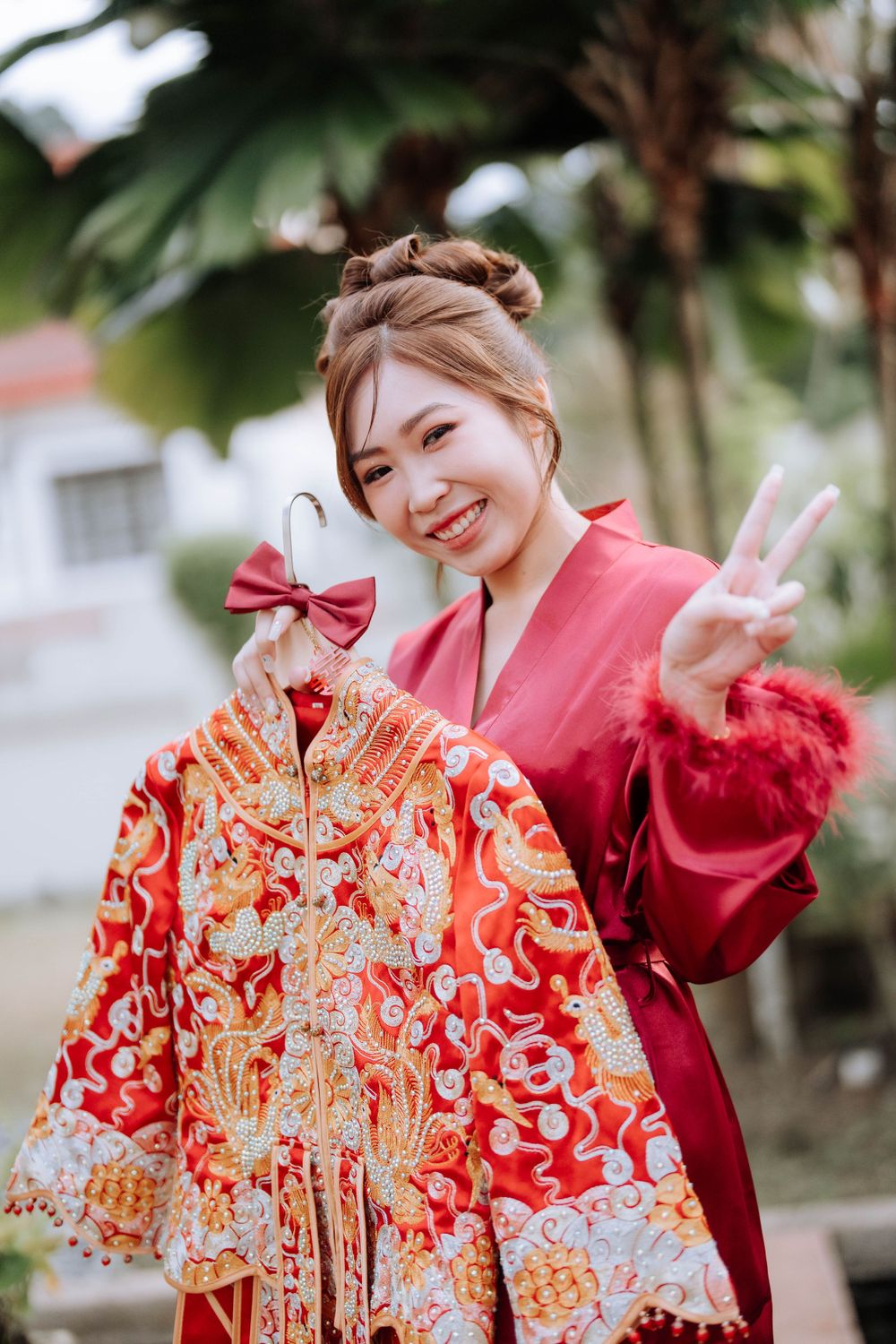 Person in red robe holding traditional Chinese wedding dress making peace sign gesture.